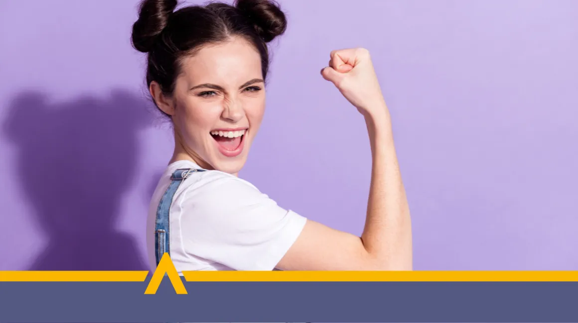 Kobieta w białej koszulce i ogrodniczkach uśmiecha się szeroko i pokazuje zaciśniętą pięść w geście zwycięstwa, na fioletowym tle.